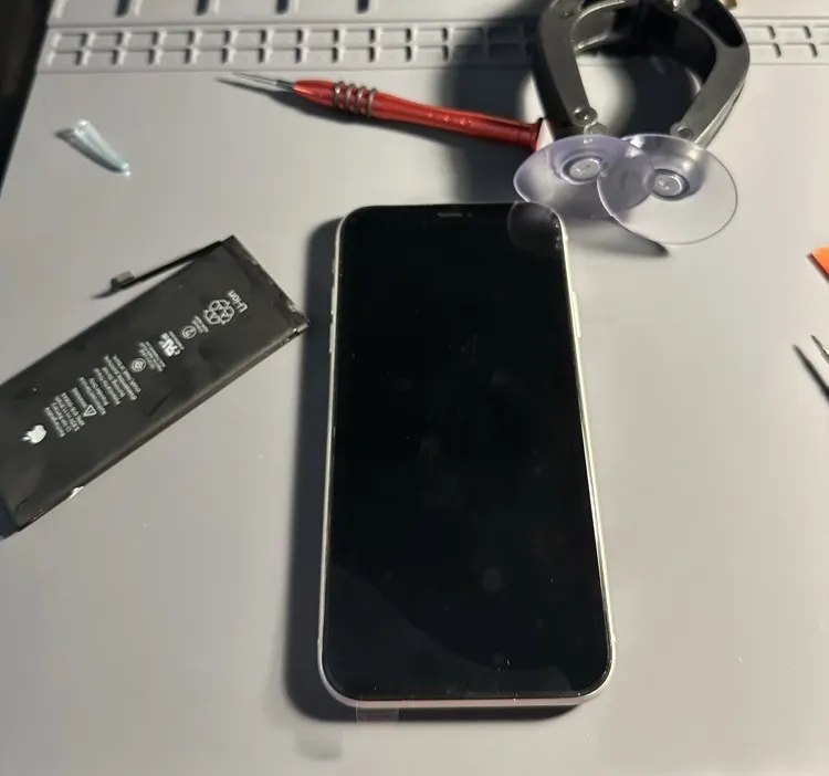 Un iPhone posé sur le tapis de l'atelier, batterie déposée à côté, tournevis de précision et ventouse à portée de main